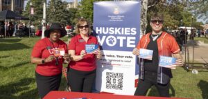 Three people stand by an information table at an outdoor event on campus. They're next to a large stand-up banner promoting the Huskies Vote initiative that includes a large QR code featuring the NIU logo.